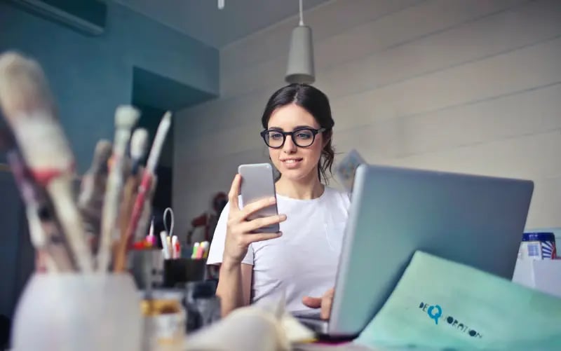 Girl looks at her phone while sitting in front of a laptop. Maybe she is checking a liveblog for a live update. 